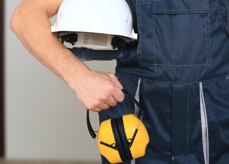 Worker holding white hard hat and safety headphones indoors. Protective wear background.